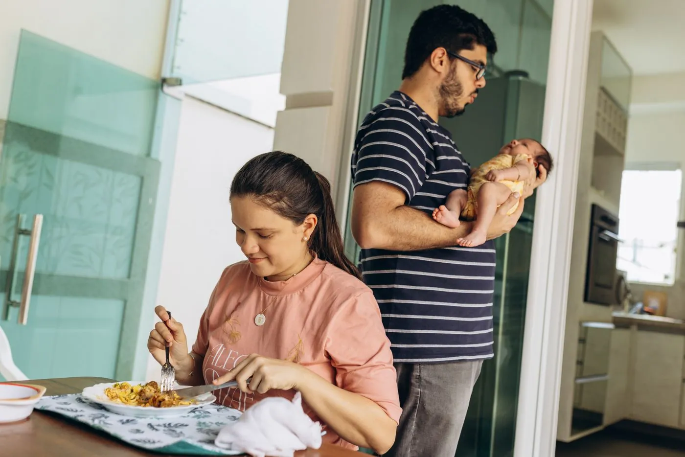 Žena jí u stolu, zatímco muž v pozadí chová miminko