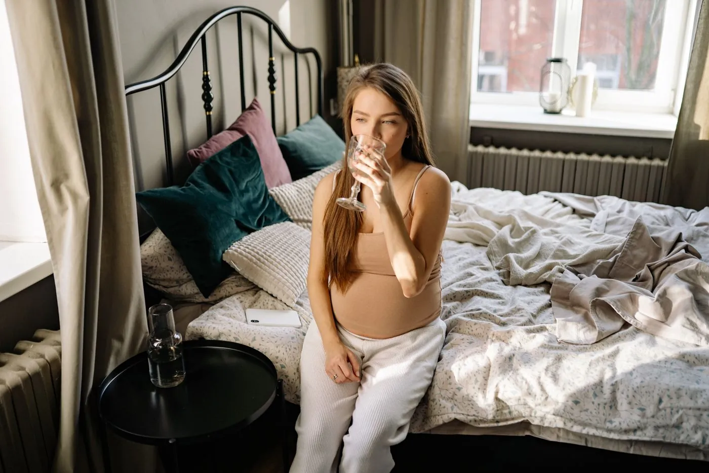 Těhotná žena sedí na posteli a pije vodu ze skla
