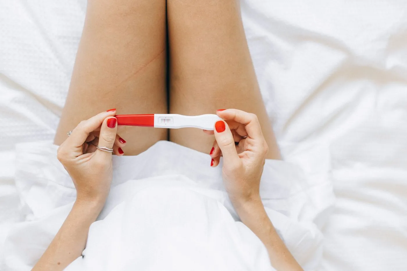 Žena leží na posteli a v ruce drží těhotenský test se dvěma čárkami